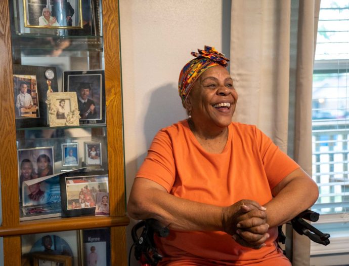 A woman sits in a chair in her home in front of a book case.