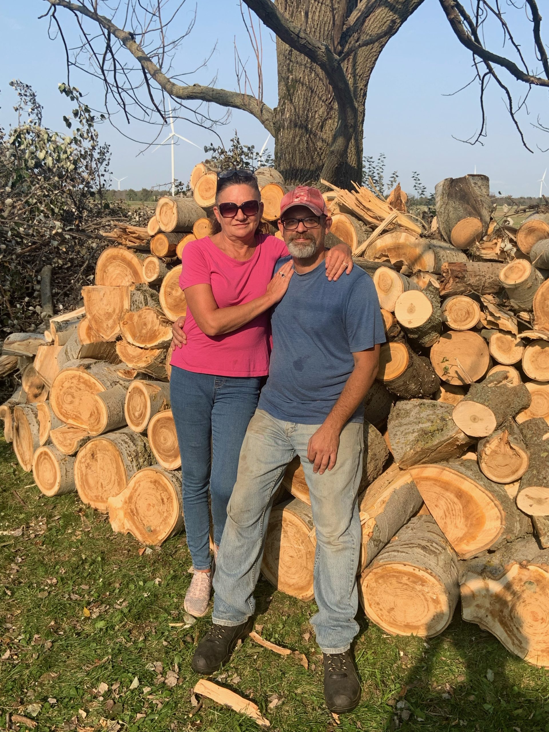 A couple stand together in front of a pile of logs.