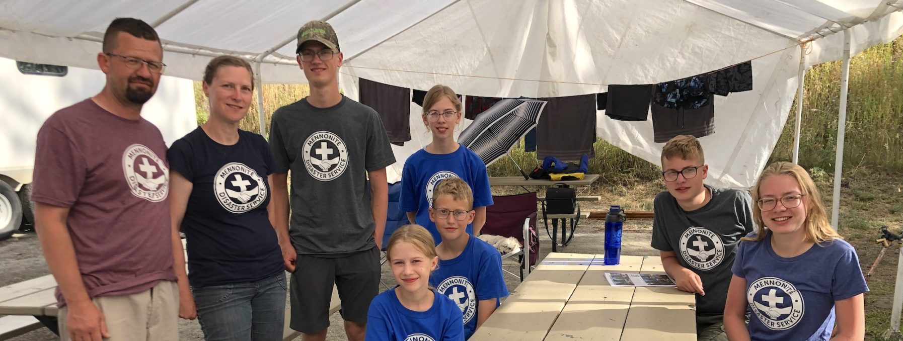 Neudorf family Peter, Susan, Joshua, emma, Jason, bigail, Andrew, Ashley pose for a picture around outdoor tables under a large tent.