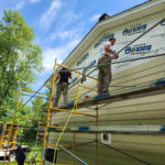 Volunteers stand on scaffolding to install tan siding on a wrapped home