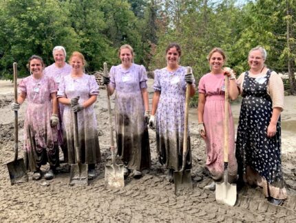 7 Traditional women stand in muddy dresses holding shovels after a long day mucking out mud.