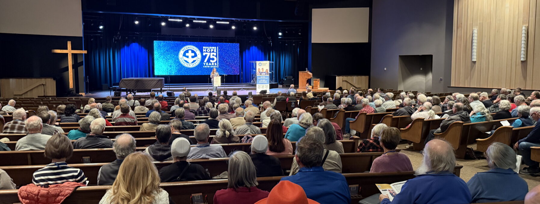 A large gathering of predominantly older adults is seated in a church or auditorium with wooden pews, attending an event. The stage features a blue-lit backdrop with a screen displaying the words "Bringing Hope 75 Years." A speaker stands at the podium, addressing the audience. A wooden cross is visible to the left of the stage. Many attendees are dressed in warm clothing, and a few are wearing distinctive hats or head coverings. Reserved seating signs for accessibility are attached to some pews.