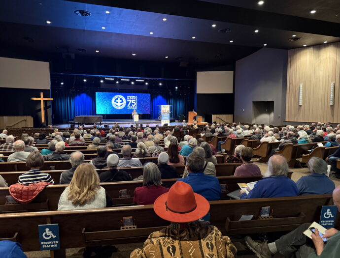 A large gathering of predominantly older adults is seated in a church or auditorium with wooden pews, attending an event. The stage features a blue-lit backdrop with a screen displaying the words "Bringing Hope 75 Years." A speaker stands at the podium, addressing the audience. A wooden cross is visible to the left of the stage. Many attendees are dressed in warm clothing, and a few are wearing distinctive hats or head coverings. Reserved seating signs for accessibility are attached to some pews.