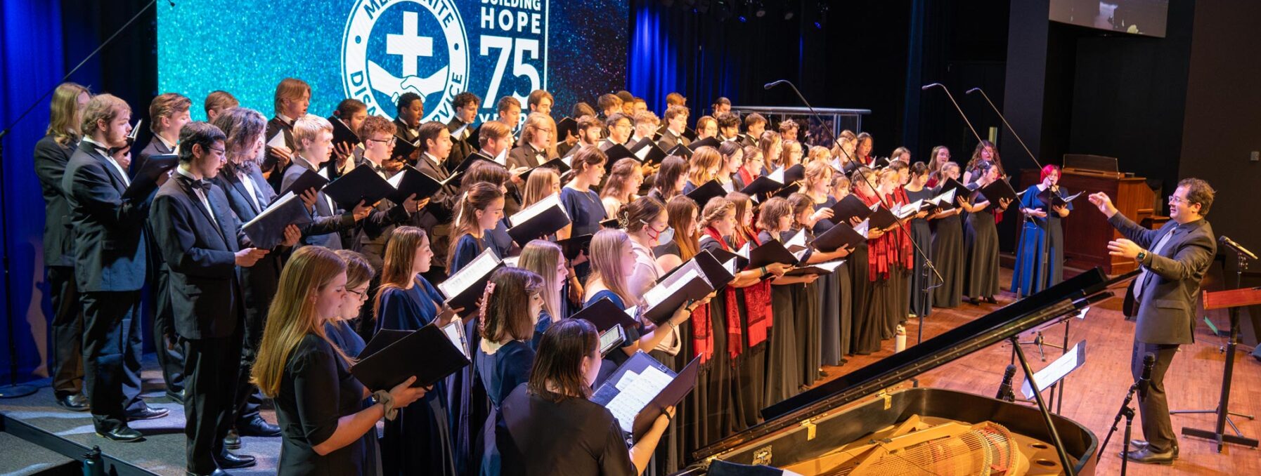 A large choir performs on stage during the Mennonite Disaster Service (MDS) 75th Anniversary Celebration. The choir, dressed in formal black attire, sings from sheet music while a conductor energetically leads them. A pianist accompanies the choir on a grand piano. The stage is illuminated with blue lighting, and a large screen in the background displays the MDS logo along with the words "Building Hope 75." A smaller screen to the side shows a live feed of the performance.