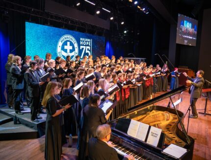 A large choir performs on stage during the Mennonite Disaster Service (MDS) 75th Anniversary Celebration. The choir, dressed in formal black attire, sings from sheet music while a conductor energetically leads them. A pianist accompanies the choir on a grand piano. The stage is illuminated with blue lighting, and a large screen in the background displays the MDS logo along with the words "Building Hope 75." A smaller screen to the side shows a live feed of the performance.