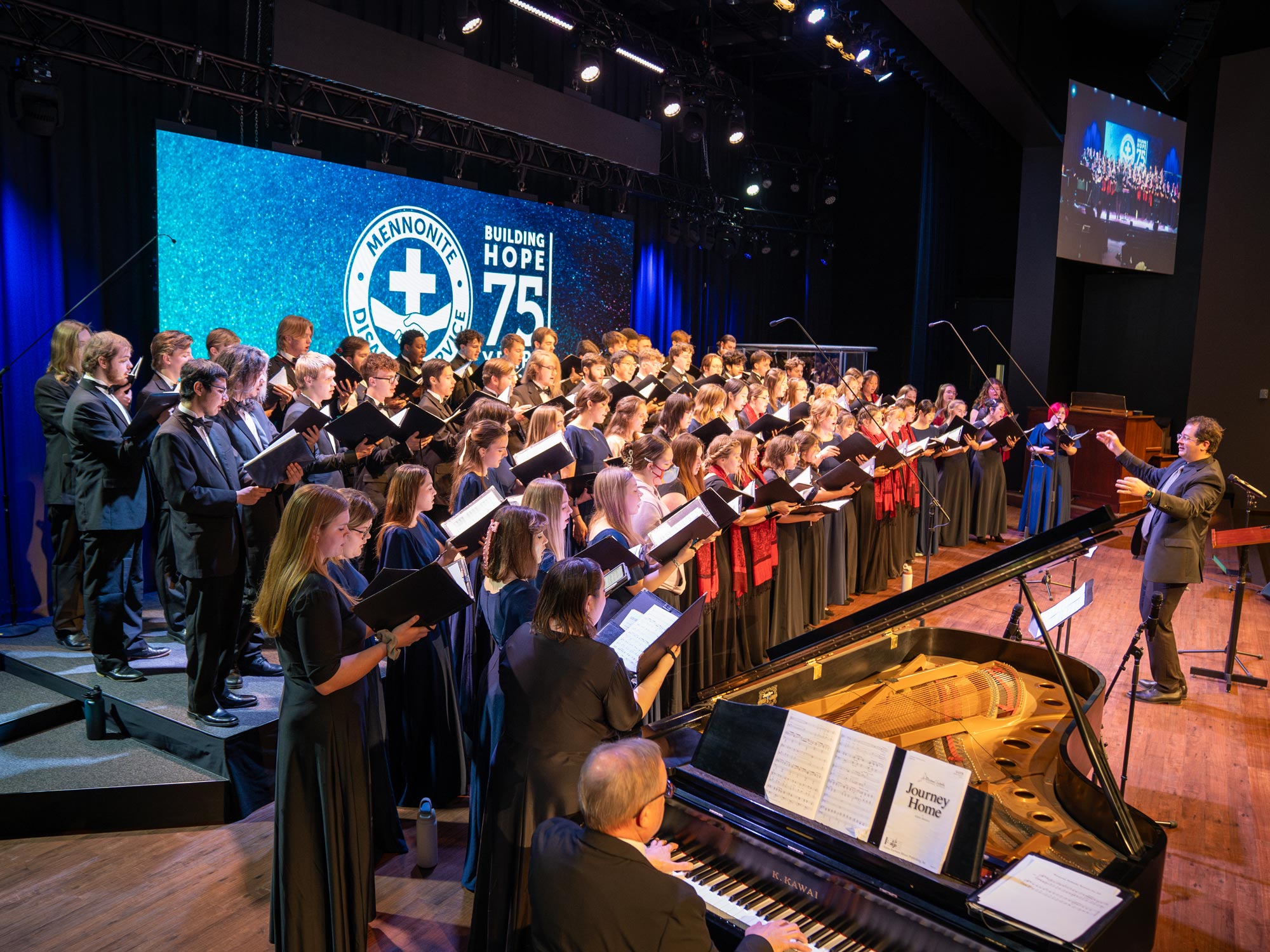 A large choir performs on stage during the Mennonite Disaster Service (MDS) 75th Anniversary Celebration. The choir, dressed in formal black attire, sings from sheet music while a conductor energetically leads them. A pianist accompanies the choir on a grand piano. The stage is illuminated with blue lighting, and a large screen in the background displays the MDS logo along with the words "Building Hope 75." A smaller screen to the side shows a live feed of the performance.