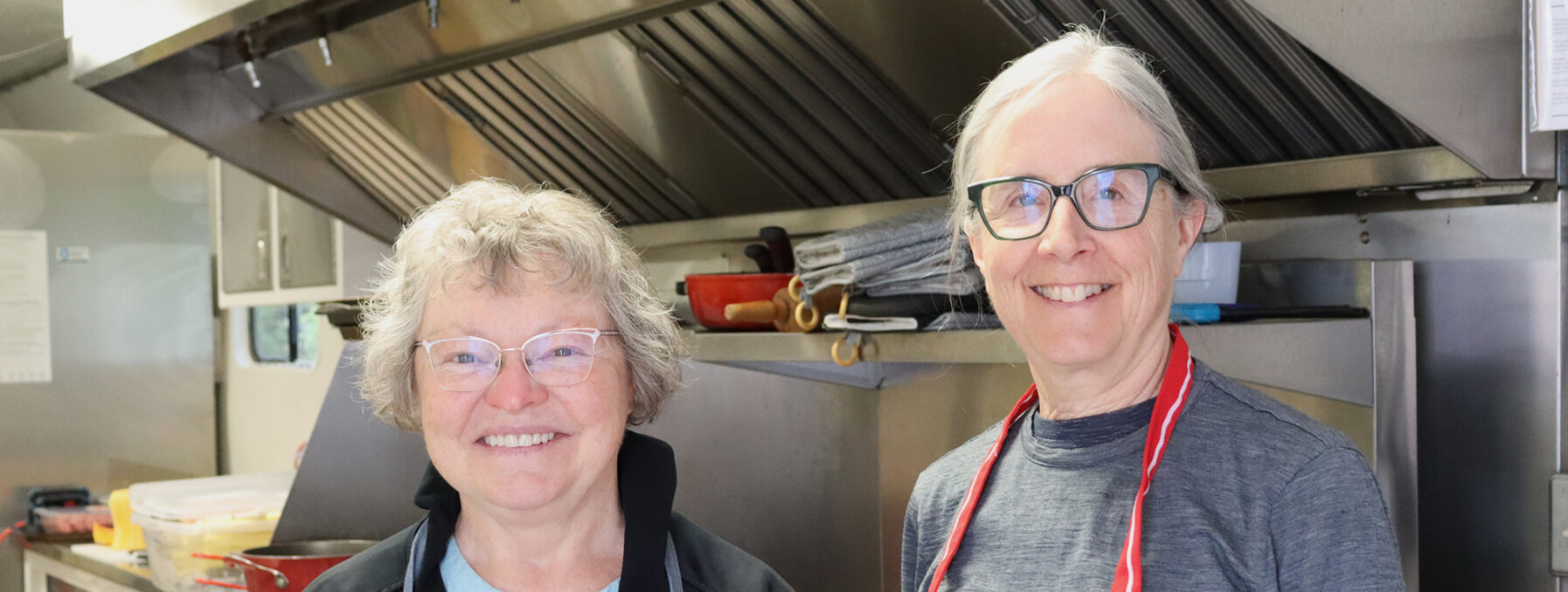 Two women, wearing aprons, stand in a kitchen