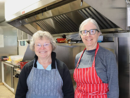Two women, wearing aprons, stand in a kitchen