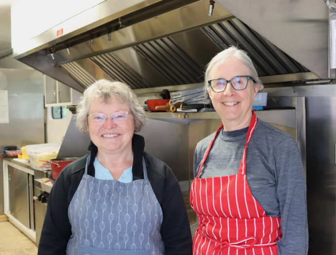Two women, wearing aprons, stand in a kitchen