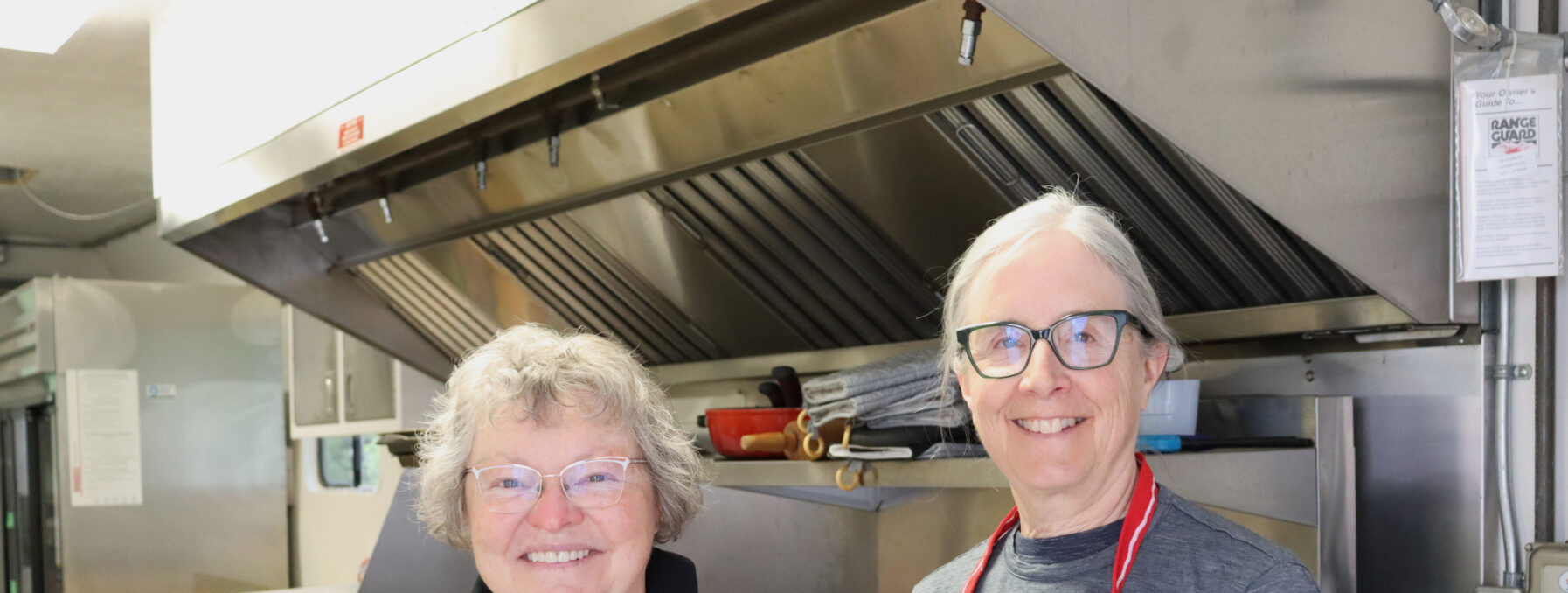 Two women, wearing aprons, stand in a kitchen