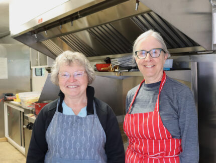 Two women, wearing aprons, stand in a kitchen