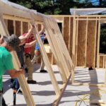 Group of men and women work togethe to raise interior framed wall.
