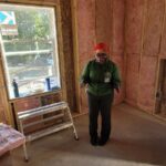 Person in green sweater stands in front of open walls of under construction home.