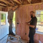 Man stands on step ladder beside man on floor, intalling insulation in walls.