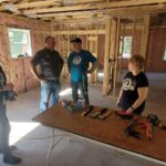 Four people stand around a table in an open house under construction.