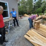 Three people stand around a pile of lumber while one of them works at fixing a small table.