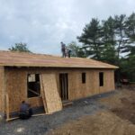 House surrounded by wooded area with three volunteers in the distance working at installing roof sheeting.