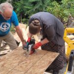 Woman uses saw to cut sheet of wood while man guides the wood.