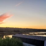 Building in the forefront with a lake, tree line, and sunrise in the background.