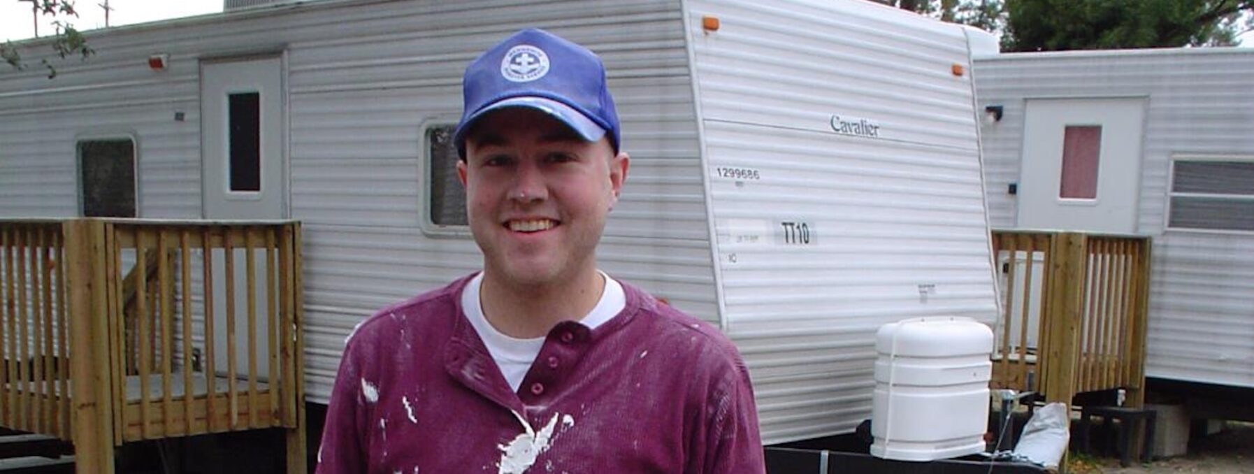 Young man in red shirt stands in front of an rv during hurricane Katrina response, he is wearing a blue MDS hat.