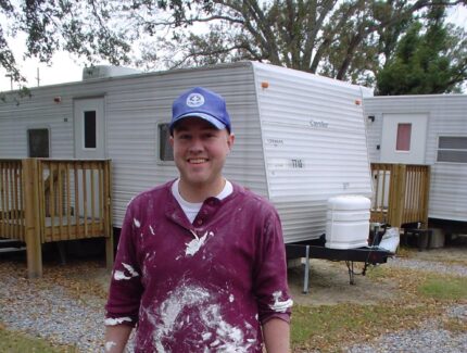 Young man in red shirt stands in front of an rv during hurricane Katrina response, he is wearing a blue MDS hat.