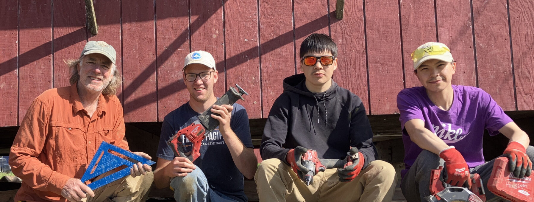 Volunteers sit in front of a red building, smiling and holding construction tools during home repair