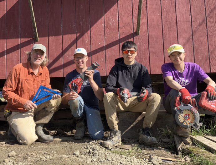 Volunteers sit in front of a red building, smiling and holding construction tools during home repair
