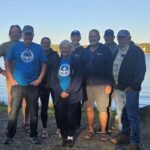 Group of 8 people stand in front of a body of water, psoing for a picture.