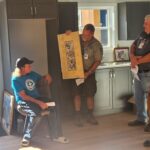 Man presents a framed picture to man sitting on chair.