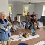 woman on left standing in a house that is under construction with two men sitting in the background holding coffee cups.