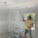 Man in yellow shirt and pants pants the ceiling of a room.