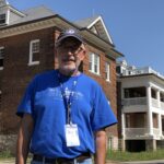 Man in blue shirt stands in front of old school building.