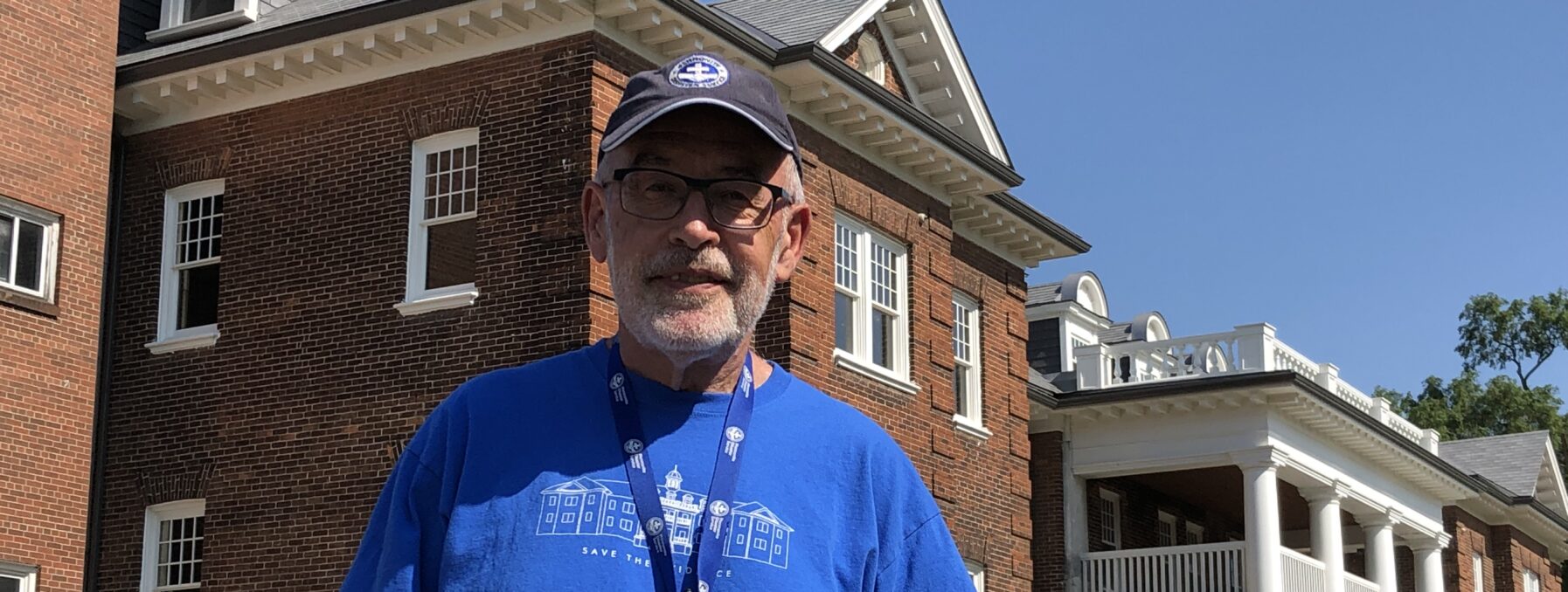 Man in blue shirt stands in front of old school building.