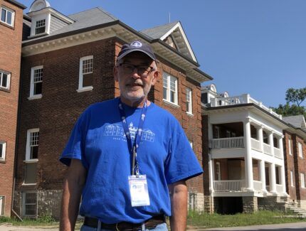 Man in blue shirt stands in front of old school building.