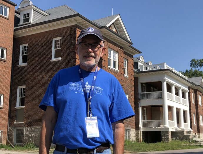 Man in blue shirt stands in front of old school building.