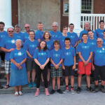 Group of people in blue shirts stand for picture outside old school building.