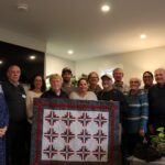 Group of people stand in a living room, holding up a quilted wall-hanging.