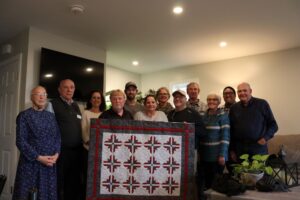 Group of people stand in a living room, holding up a quilted wall-hanging.