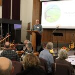 Man speaks from podium on stage in front of seated crowd.