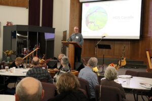 Man speaks from podium on stage in front of seated crowd.