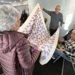 Two people, standing, hold up a quilt in front of two people, sitting.