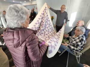 Two people, standing, hold up a quilt in front of two people, sitting.