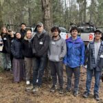 Group of people standing together in a forest, in front of vehicles, posing for picture.