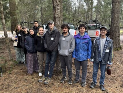Group of people standing together in a forest, in front of vehicles, posing for picture.