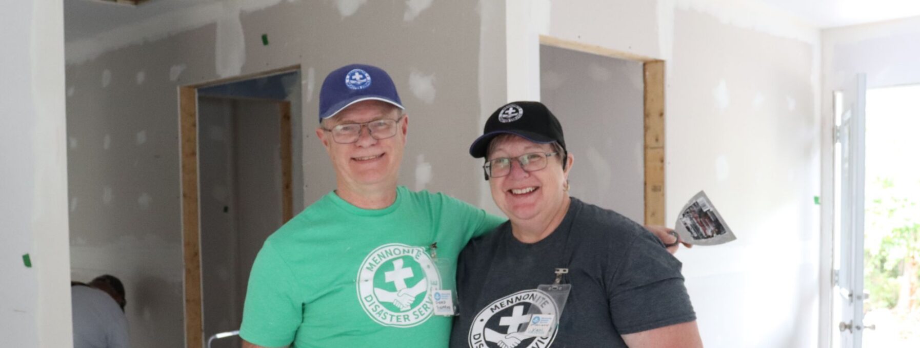 Two people standing in a home under construction holding mudding supplies.