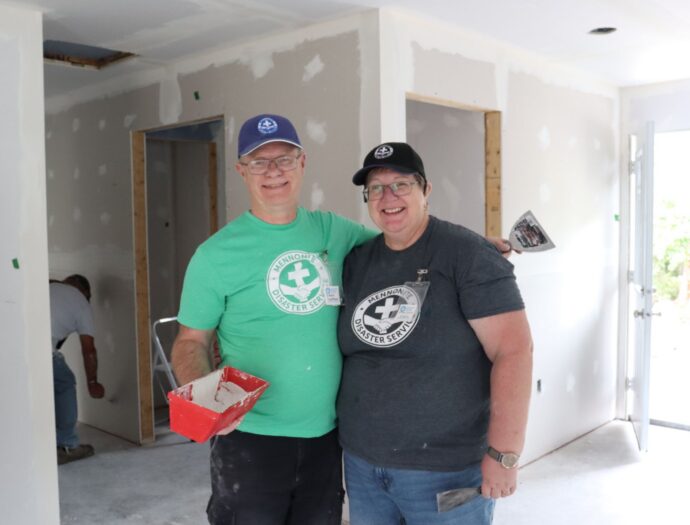 Two people standing in a home under construction holding mudding supplies.