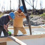 Two men working on pouring foundations, one with a trowel and the other guiding the cement pipe.
