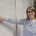 Woman stands beside a map, pointing to a spot on it.