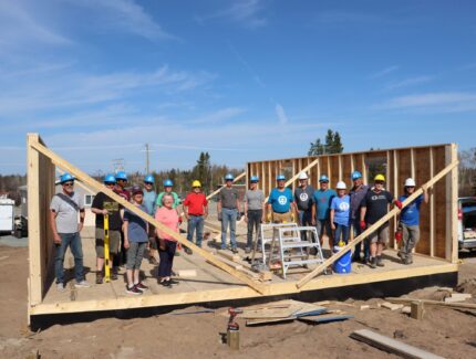 Group of people stand, posing for picture, inside a half-framed house.